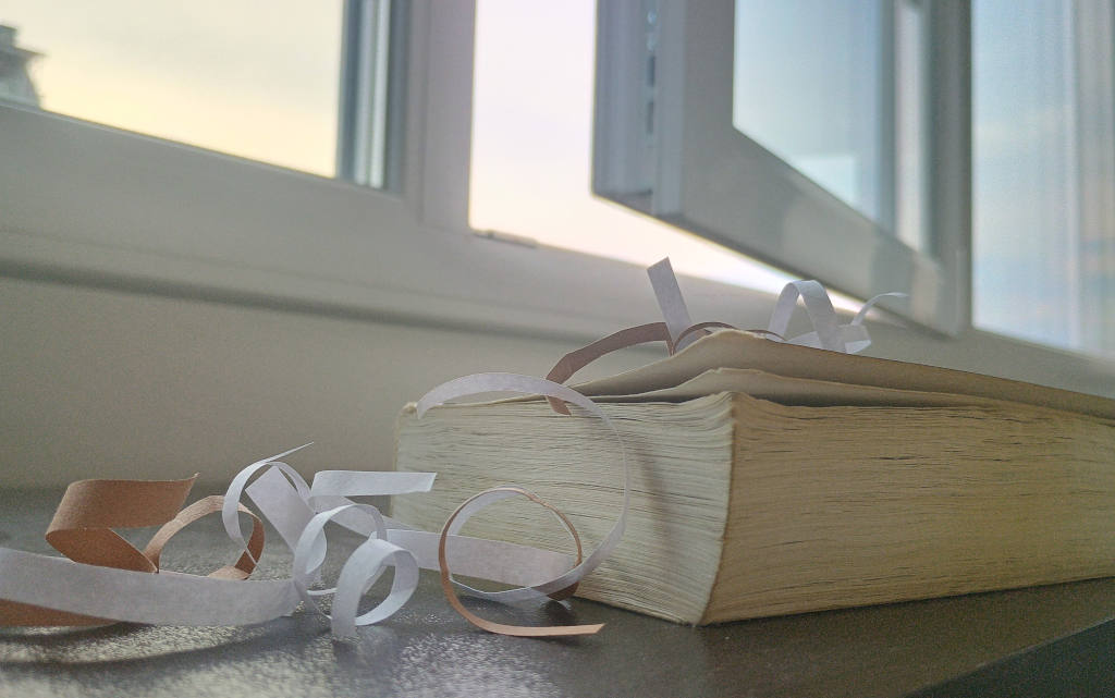 ^Abstract photograph of a yellowed book surrounded by a few paper streamers in front of an open window