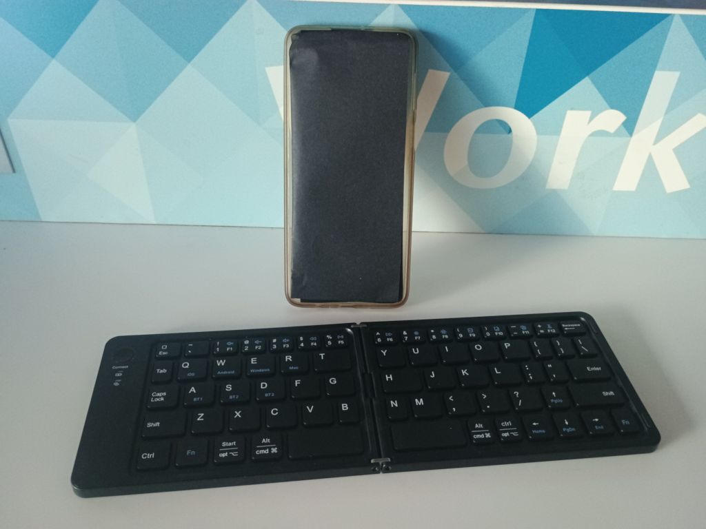 Photo of the foldable keyboard unfolded next a phone (mocked out by a piece of folded black paper) at a random "Work" table at the airport