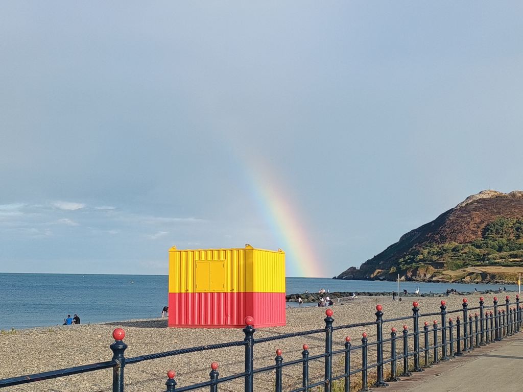A rainbow over the beach at Bray, Ireland, with a red-yellow container in front